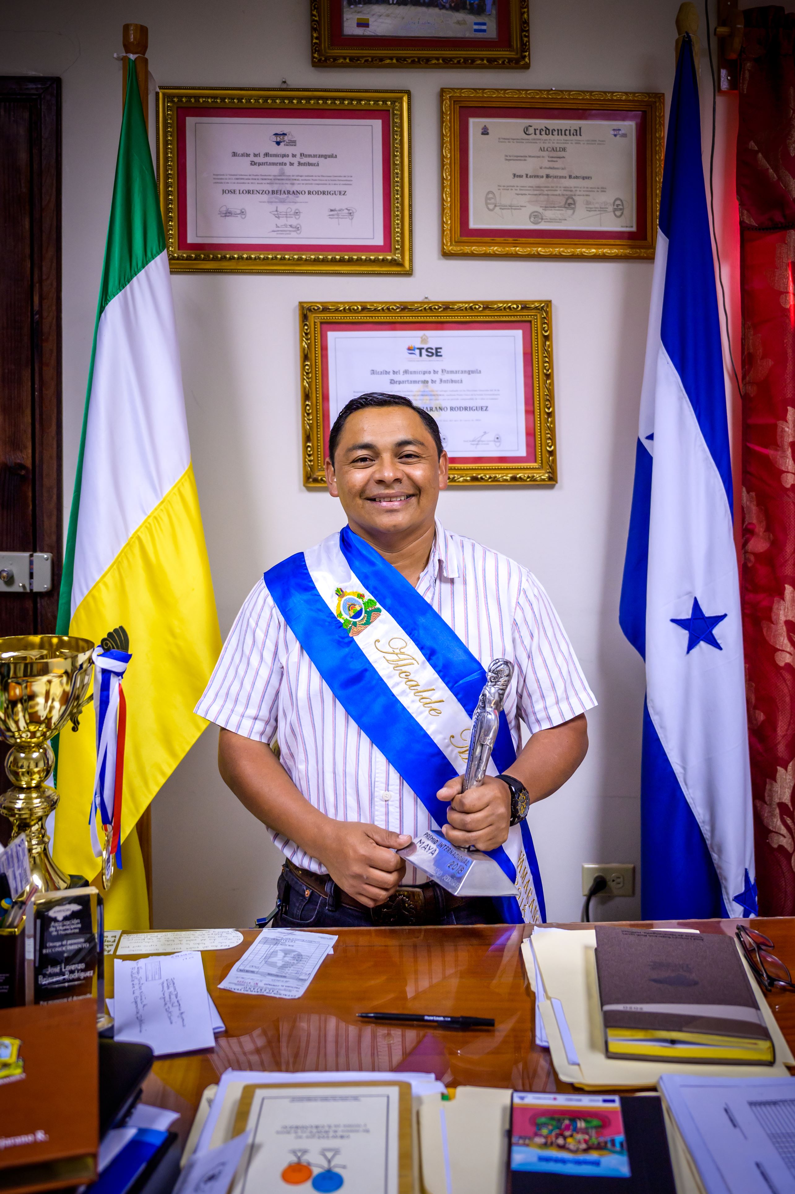 Hombre de Honduras que lleva una banda y sostiene un trofeo mientras est&aacute; de pie en su oficina entre dos banderas